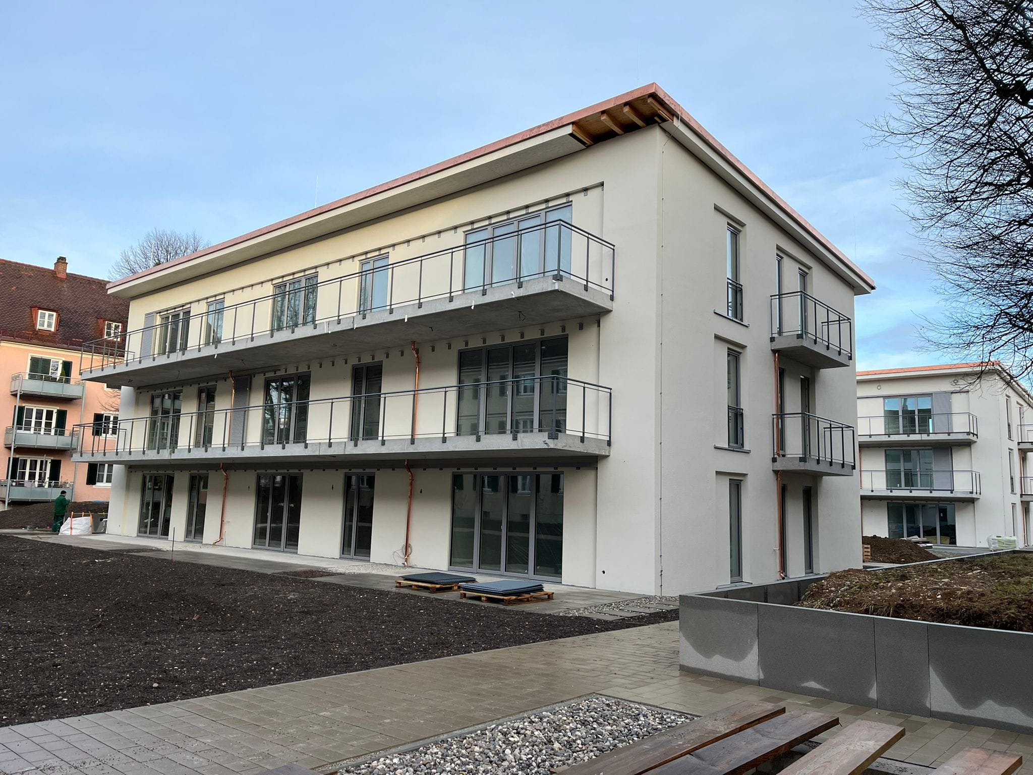 Ein moderner Neubau eines Mehrfamilienhauses in der Endphase der Bauarbeiten. Das Foto zeigt die helle, fertig verputzte Fassade, eingesetzte dunkelgraue Fenster und durchgehende Balkone, die noch den rohen Betonboden aufweisen. Im Vordergrund dominiert die unfertige Außenanlage mit dunklem Erdboden und begonnenen Pflasterarbeiten.